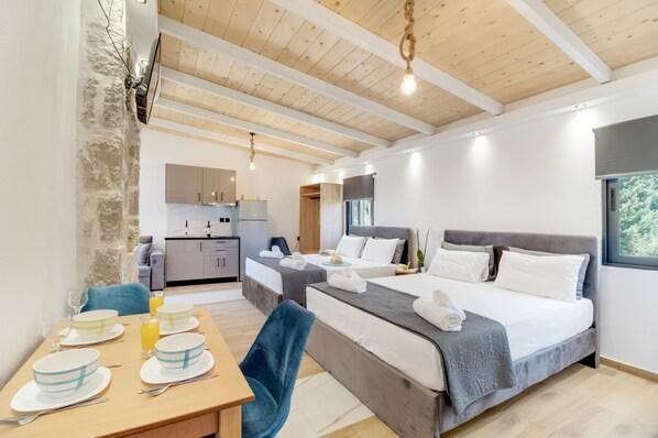 A modern hotel room with a large bed, a dining area with a table set for two, a kitchen in the background, and a high wooden ceiling.