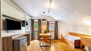 A modern, compact room with a wooden dining table, a bed, a built-in TV, and a small kitchenette. - Residence Grünbacher Apartment 4 with Balcony and Shared Pool (Issengo)