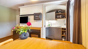 A modern room with a wooden dining table, a vase with flowers, a flat-screen TV, a kitchen area with cabinets and a microwave, and a large window with curtains. - Residence Grünbacher Apartment 4 with Balcony and Shared Pool (Issengo)