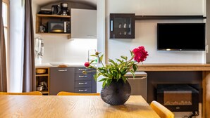 Un comedor con mesa y sillas de madera, un florero con flores, una cocina al fondo y un televisor de pantalla plana montado en la pared.