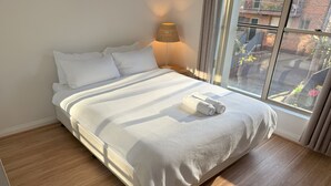 Ein ordentlich bezogenes Bett mit weißer Decke und Kissen, ein zusammengerolltes Handtuch und eine Lampe auf einem Nachttisch.