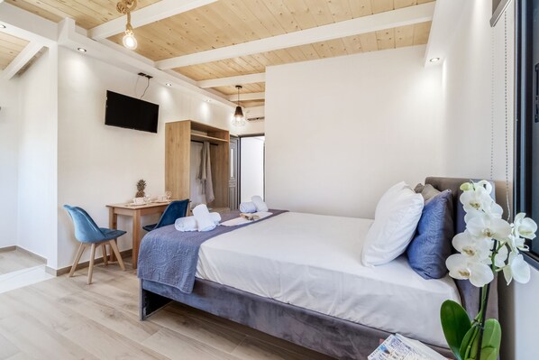 Un dormitorio moderno con una cama, un escritorio, una silla, un televisor y una ventana adornada con orquídeas blancas.