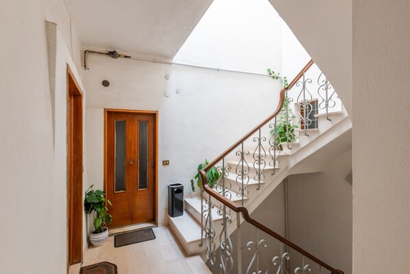 A narrow hallway with a wooden door, a staircase with metal railings, and potted plants.