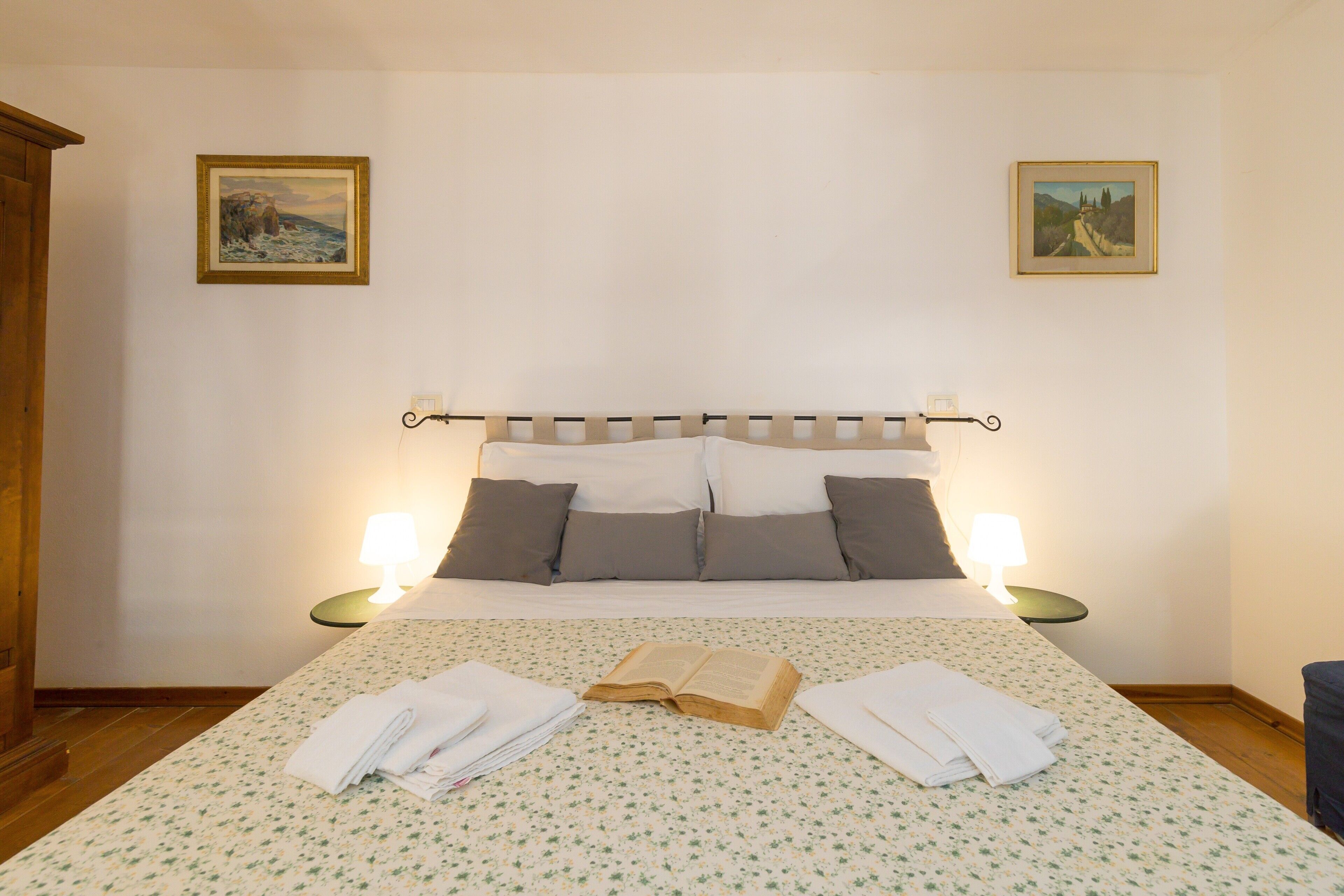 Een net opgemaakt bed met een bloemenbedekker, twee bedlampen, een boek en handdoeken op het bed.