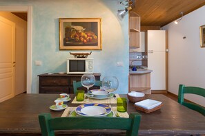 Un coin repas avec une table en bois, des chaises vertes, un tableau représentant des fruits accroché au mur, un four à micro-ondes et un réfrigérateur.