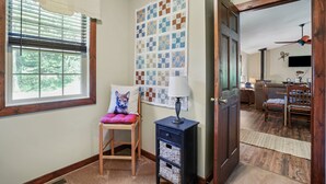 Ein Zimmer mit Fenster, ein Sessel mit Kissen, eine Lampe, eine Wand mit einer Quilttdecke und eine Tür zu einem anderen Raum.