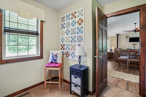 A room with a window, a chair with a cushion, a lamp, a wall with a quilt, and a door leading to another room.