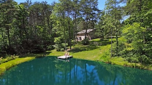 Un lac paisible, avec un quai et des chaises, entouré d’une végétation luxuriante et une maison en arrière-plan.
