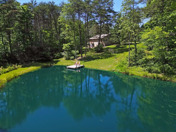 Un lago tranquillo con un molo e sedie, circondato da vegetazione lussureggiante e una casa sullo sfondo.