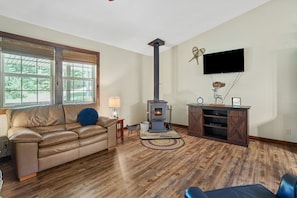 A living room with a leather sofa, a wood-burning stove, a TV mounted on the wall, and a wooden floor. - Boondocks Bungalow | Hocking Hills, 18 Acres, Pond (New Plymouth)