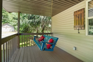 Un porche abrité avec un banc bleu et des coussins rouges, une balustrade en bois et une fenêtre dotée d’un volet en bois.