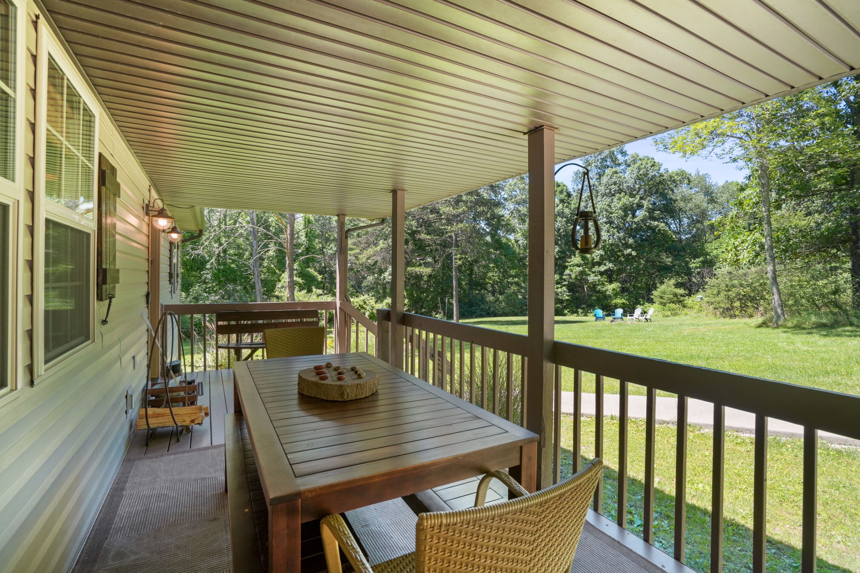 A covered porch with a wooden table and chairs, a small table with a basket, and a view of a grassy yard with trees.