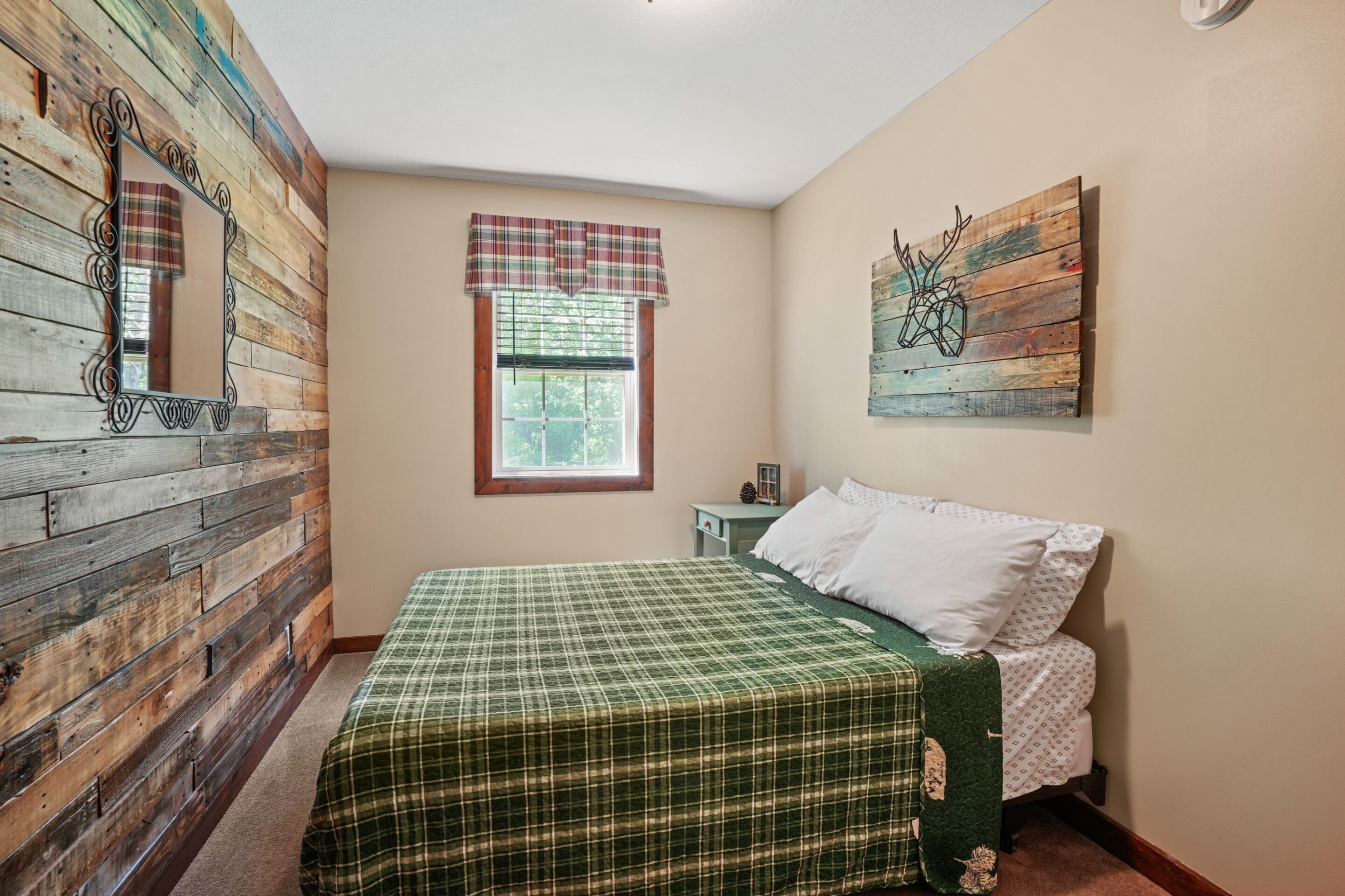 A bedroom with a wooden wall, a window with a plaid valance, a bed with a green checkered bedspread, and a wall-mounted artwork.