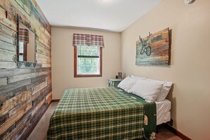 Un dormitorio con pared de madera, una ventana con cortina escocesa, una cama con colcha verde a cuadros y una obra de arte colgada en la pared.