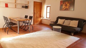 Una sala de estar con un sofá, una mesa de comedor con sillas, una mesita redonda, un tapete blanco, una puerta de madera, una ventana y un cuadro colgado en la pared.