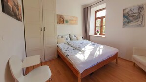 Un dormitorio con cama, una silla, armario, ventana y obras de arte en las paredes.