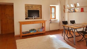 Una habitación con una televisión sobre un soporte de madera, una mesa con sillas, un tapete blanco, una ventana con cortinas y una estantería con libros y un letrero rojo.