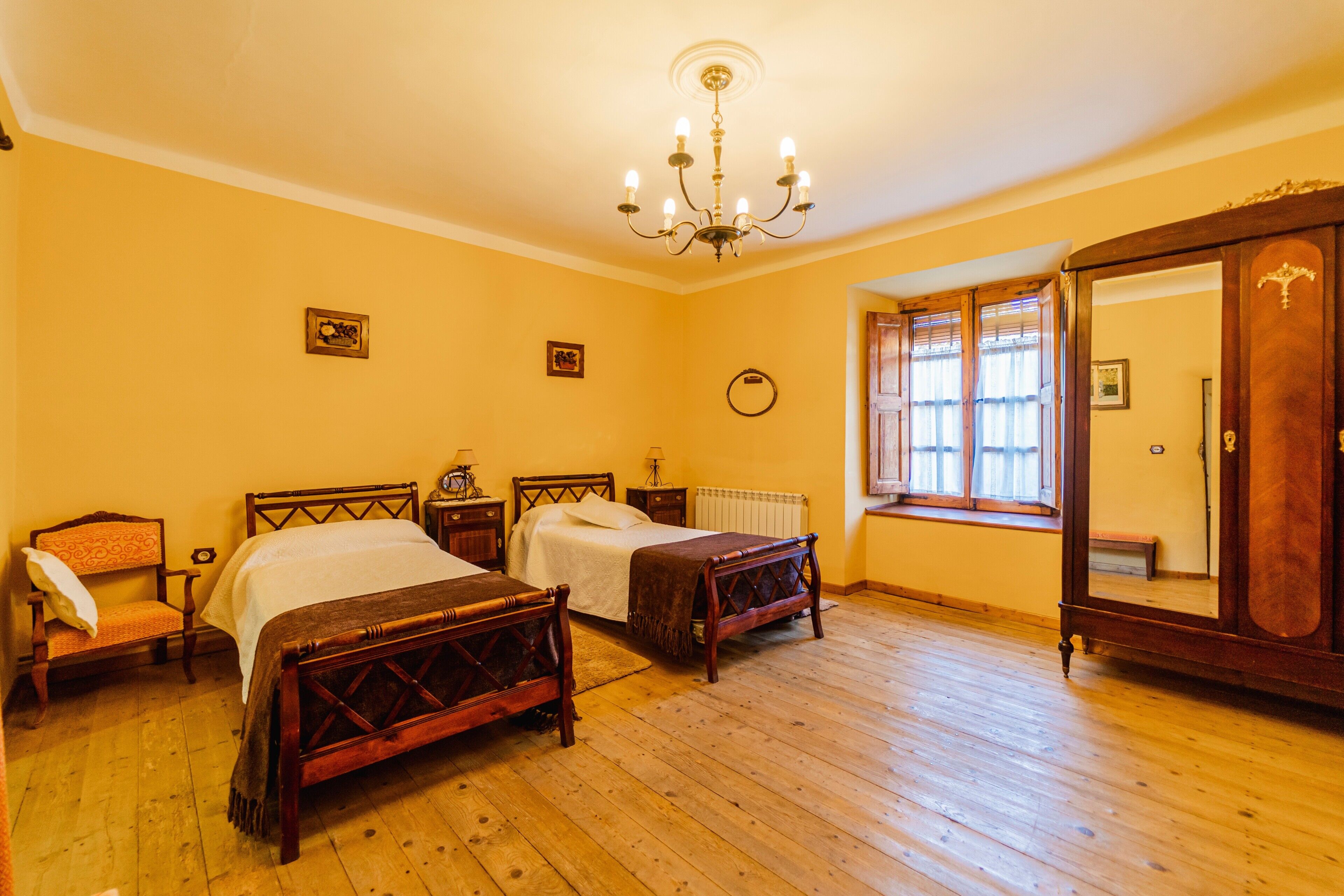 A bedroom with two wooden beds, a wooden wardrobe, a window with curtains, and a chandelier.