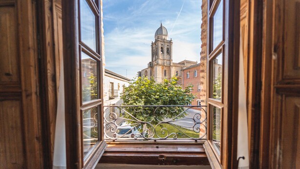 An open window with a view of a building with a bell tower, a metal railing, and a tree.