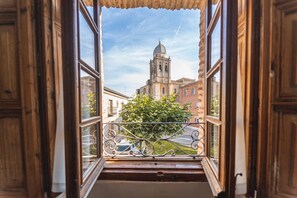 Una ventana abierta con vistas a un edificio que tiene un campanario, una barandilla metálica y un árbol.