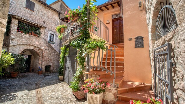 Une rue pavée, avec un bâtiment en pierre, un escalier en bois et des plantes en pot.