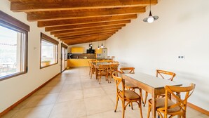 A spacious dining area with wooden chairs and a long table, a kitchen in the background, and large windows along the walls.