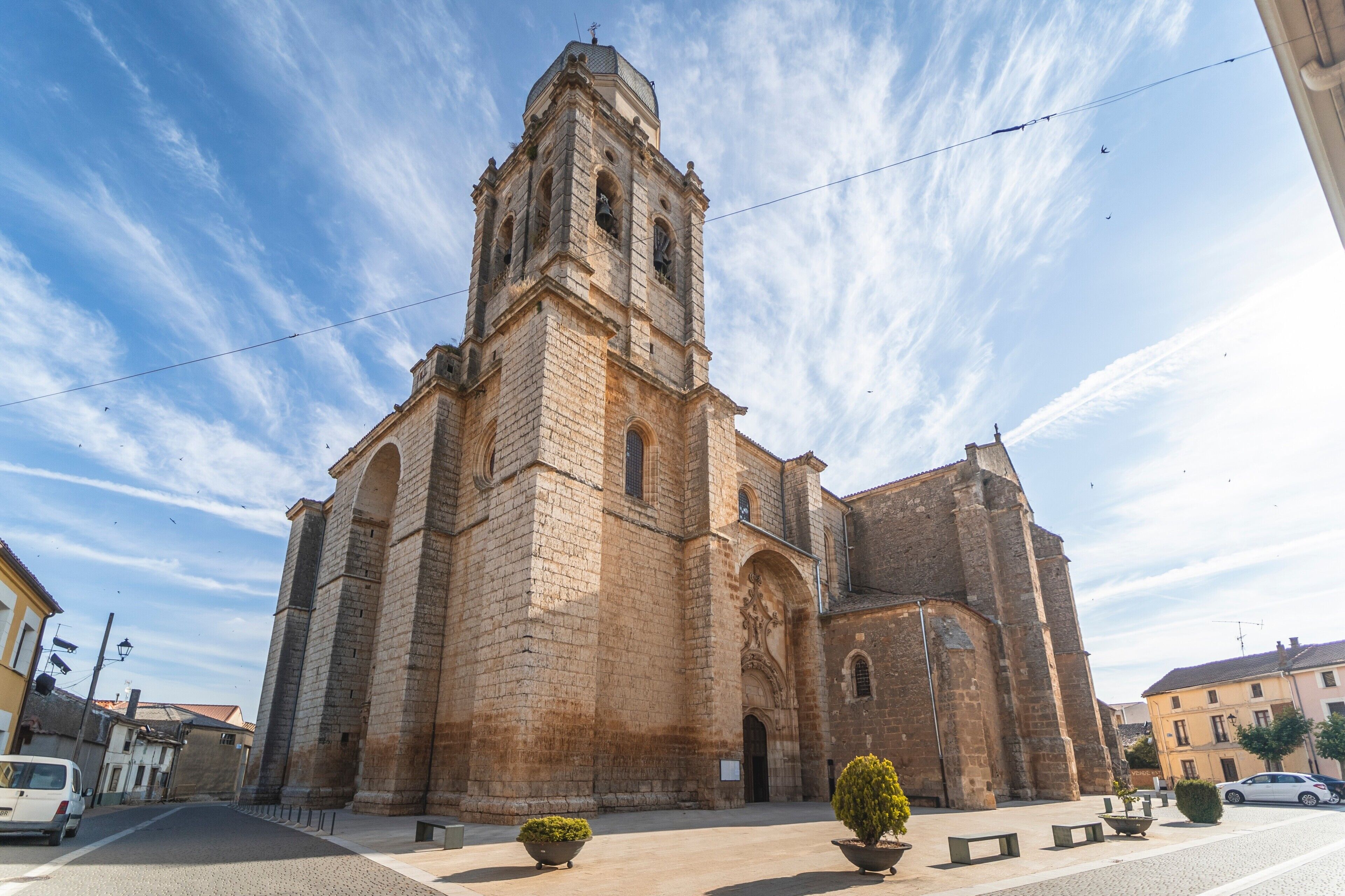 Una iglesia de piedra histórica con un campanario destacado, un patio empedrado y un cielo despejado.