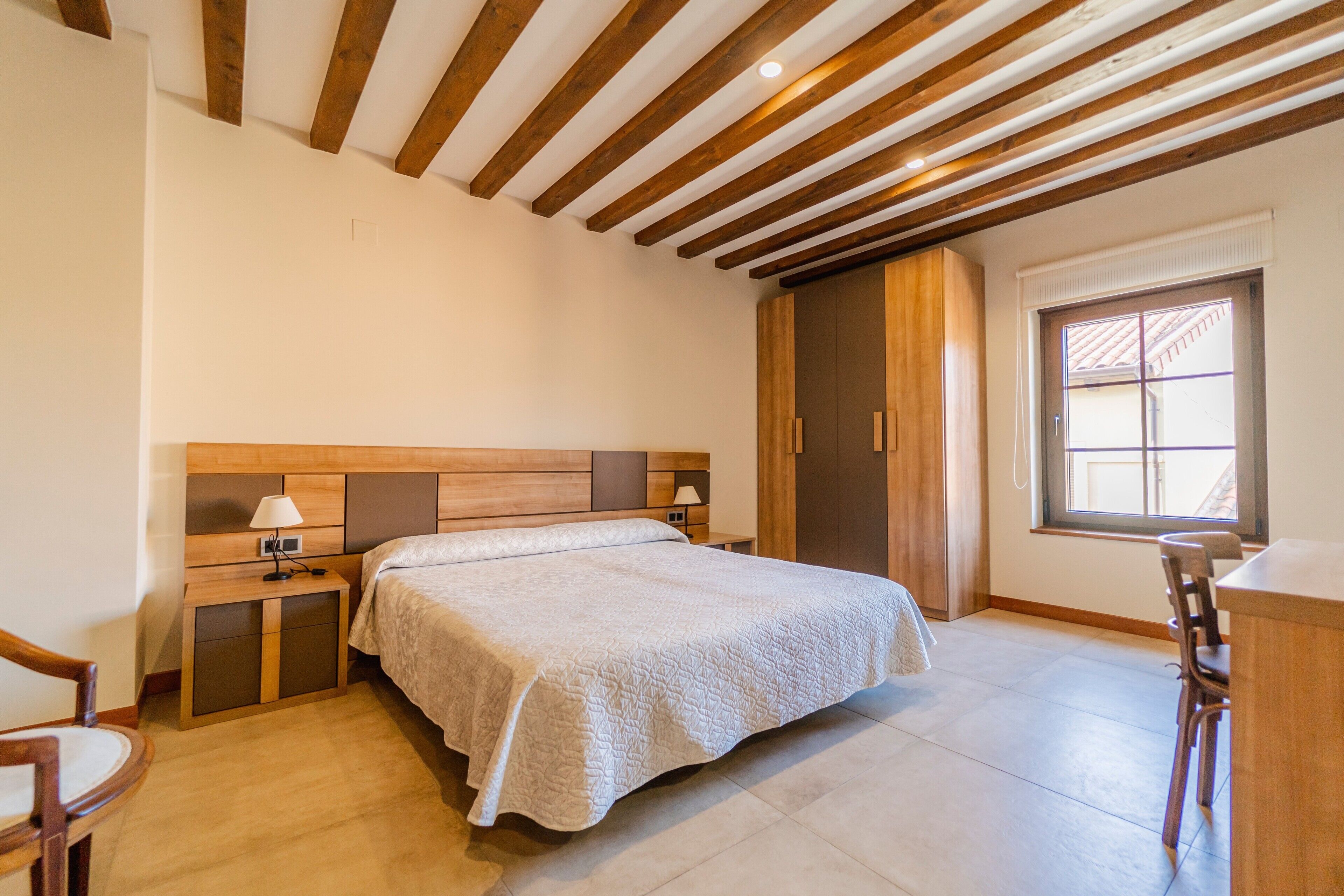 Ein Schlafzimmer mit Holzbalkendecke, einem Bett mit weißer Bettdecke, einem Nachttisch mit Lampe und einem Fenster mit Blick.