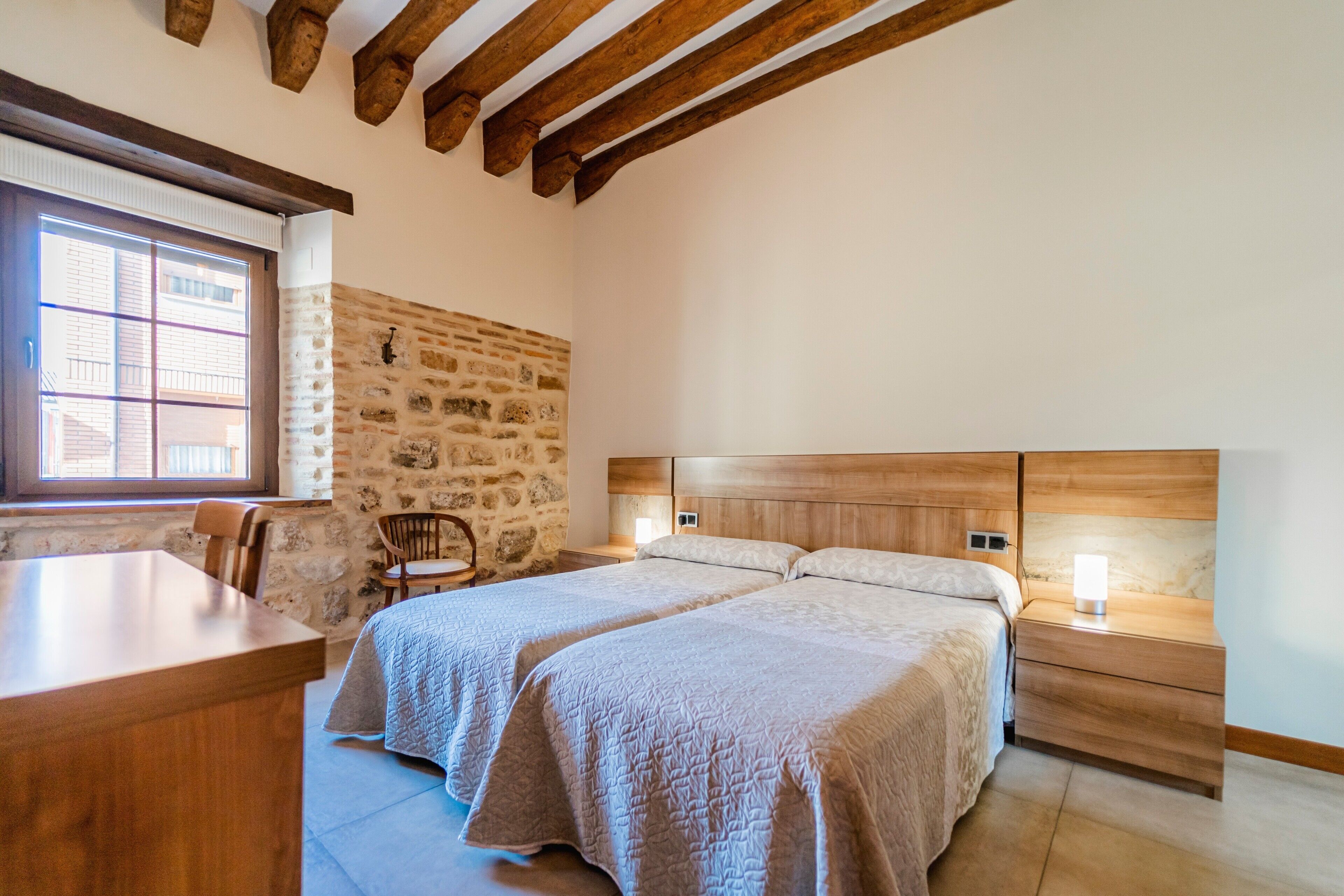 Un dormitorio con dos camas, un escritorio de madera, una pared de piedra y una ventana con persianas.