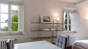 Ein Zimmer mit weißem Tisch, einem Sessel, einem Bett, einem Heizkörper und einem Fenster mit Blick auf Bäume.