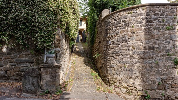 Ein schmaler, gepflasterter Weg, der von Mauern gesäumt wird und zu einem Gebäude mit Bogenöffnung führt.