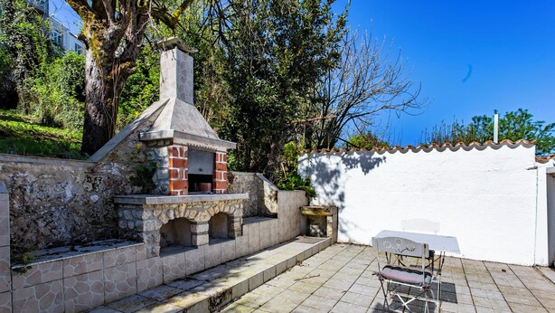 A patio with a stone barbecue, a white wall, and a white table with a chair.