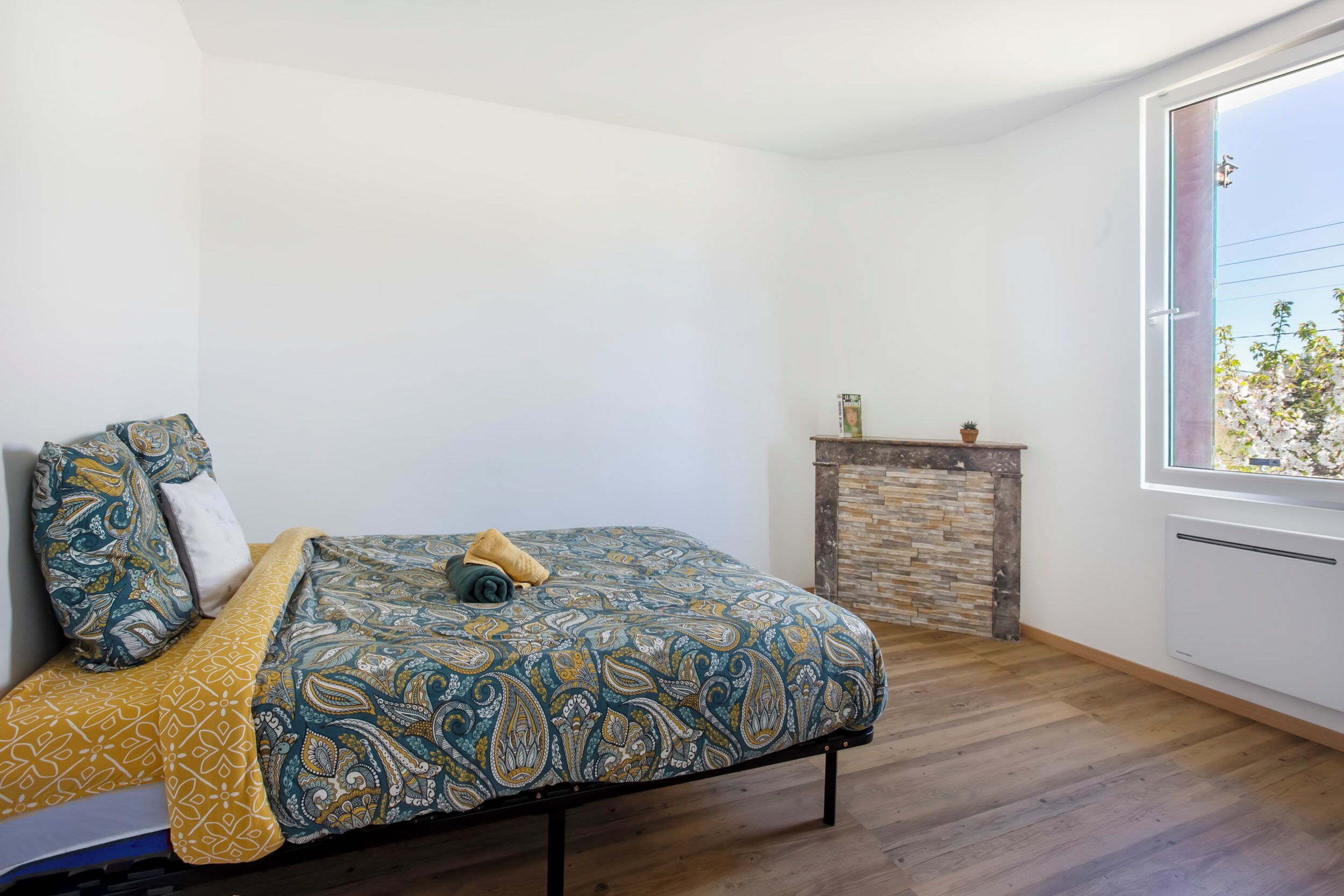 A bedroom with a bed, a small fireplace, and a window with a view of trees.