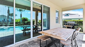 Patio con tavolo e sedie di legno, barbecue e vista su piscina e vegetazione.