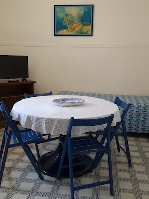 A round table with a white tablecloth and blue chairs, a painting on the wall, and a television on a wooden stand. - Villa 'La Pineta' with Shared Garden and Wi-Fi (Favignana)