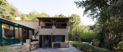 A modern two-story house with a stone patio, a covered outdoor seating area, and a view of the surrounding forest.