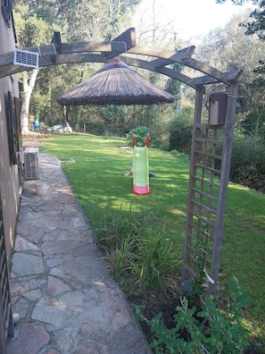 Un camino de piedra que conduce a una pérgola de madera con un toldo de paja, una silla verde y un comedero para pájaros.