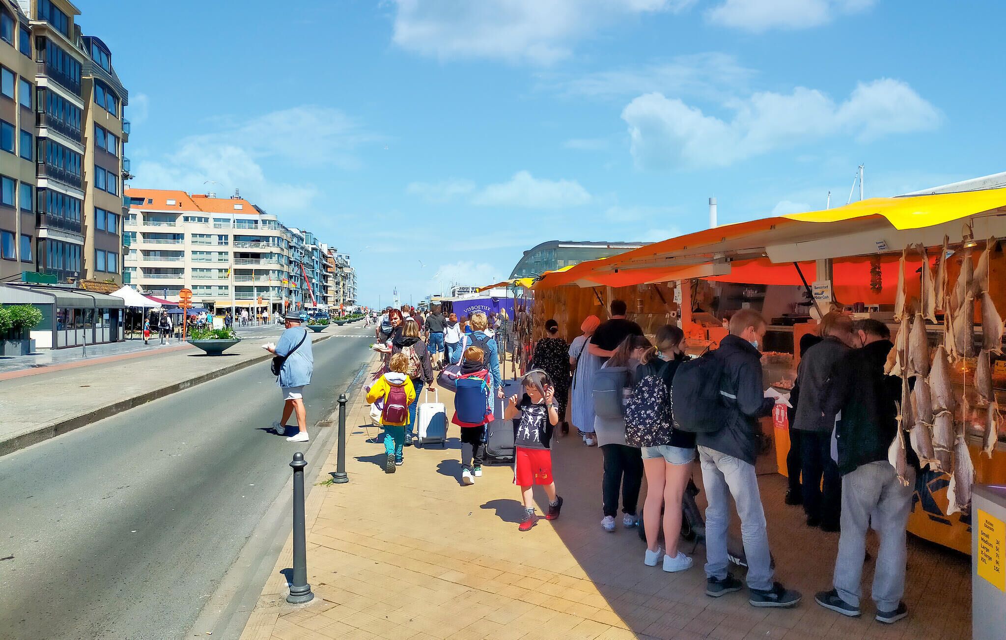A bustling street with people shopping at outdoor stalls, a clear sky, and modern buildings in the background.