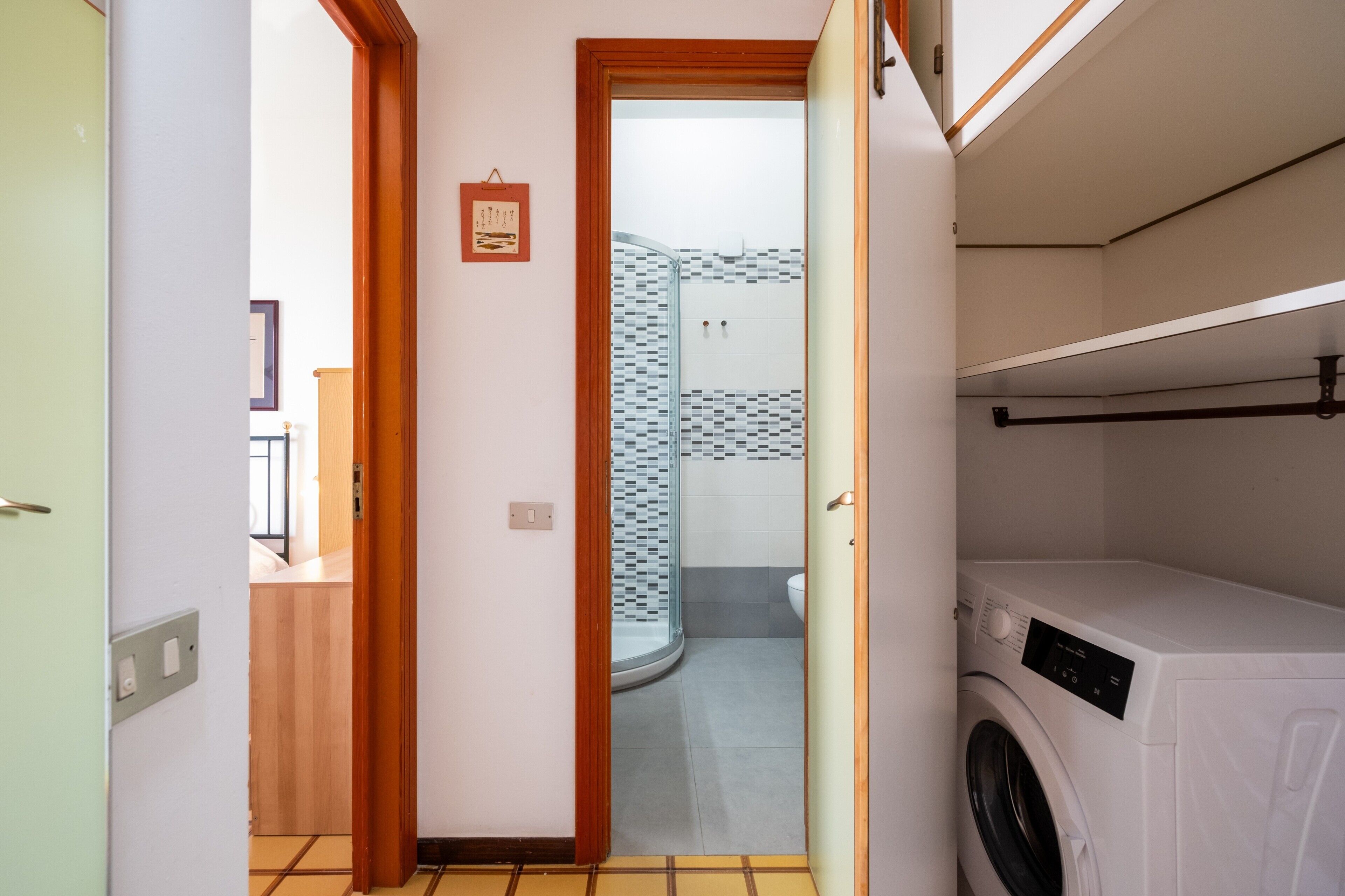 Un área de lavandería con lavadora, una ducha con paredes de azulejos y una puerta de madera que da a otra habitación.