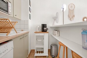 Uma cozinha compacta com micro-ondas, cafeteira e torradeira na bancada, um carrinho pequeno com suporte para vinhos e uma lixeira.