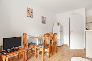 Una habitación pequeña con televisión, sillas de madera, una mesa, un refrigerador y dos cuadros en la pared.