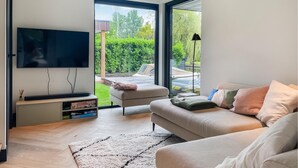 Een moderne woonkamer met een flatscreen-tv, een bank en een salontafel. Door de glazen deuren is er uitzicht op een tuin en een auto.