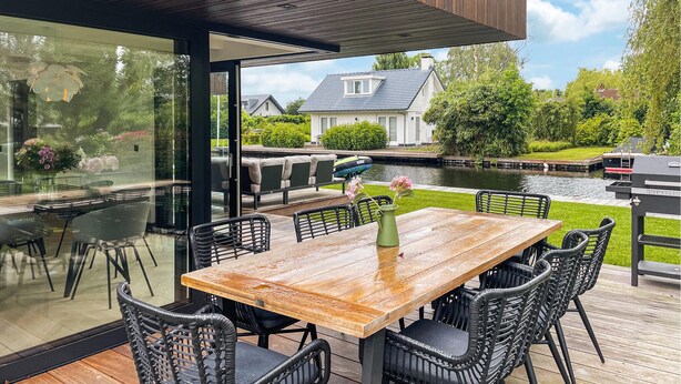Een modern buitenmeubelgedeelte met een houten tafel en zwarte rieten stoelen, met uitzicht op een gracht met een huis en groen op de achtergrond.