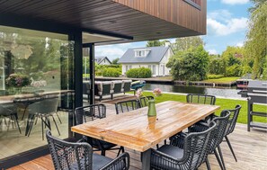 Uma área de refeições moderna ao ar livre, com mesa de madeira e cadeiras de vime preto, com vista para um canal, com uma casa e vegetação ao fundo.