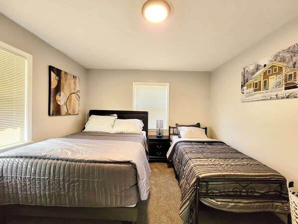 A bedroom with two beds, a nightstand, a wall-mounted artwork, and a window with blinds.