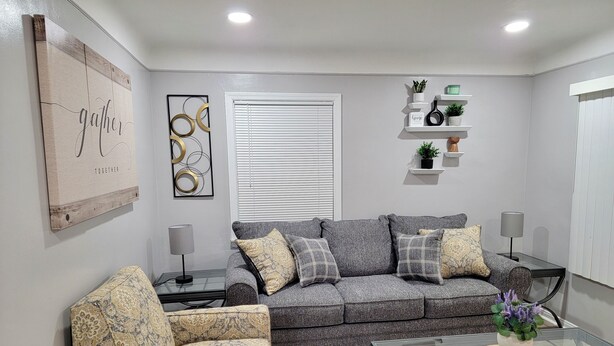 Ein Wohnzimmer mit grauem Sofa, einem gemusterten Sessel, einem Glastisch und Dekorationen auf Regalen.