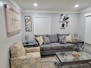 A living room with a grey sofa, patterned armchair, glass coffee table, and decorative items on shelves.