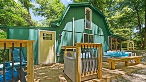 Eine grüne Holz-Blockhütte mit Veranda, einer Tür und einem Fenster. Auf der Veranda stehen ein blauer Stuhl und ein Korbstuhl.