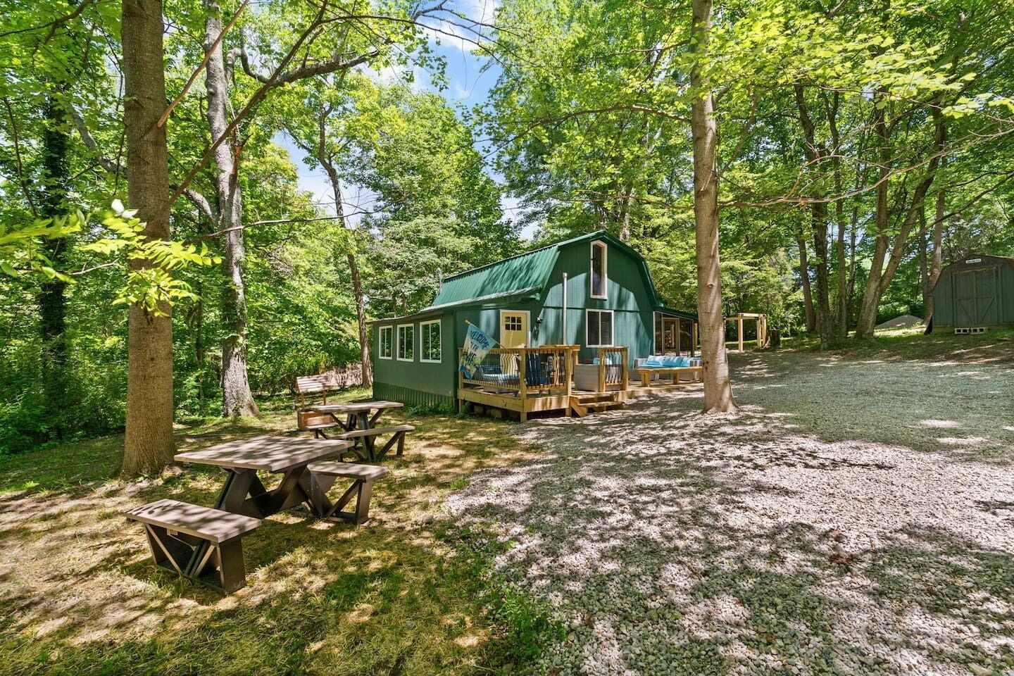 Een rustieke houten hut met een groen dak, omgeven door bomen, met picknicktafels en een terras met zitplaatsen.