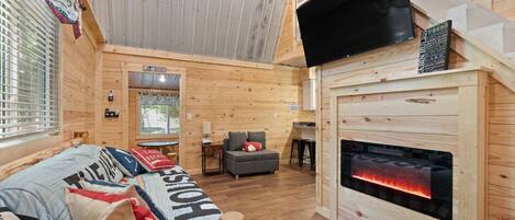 A cozy cabin-style room with a bed, a sofa, a fireplace, a television, and a window with blinds.